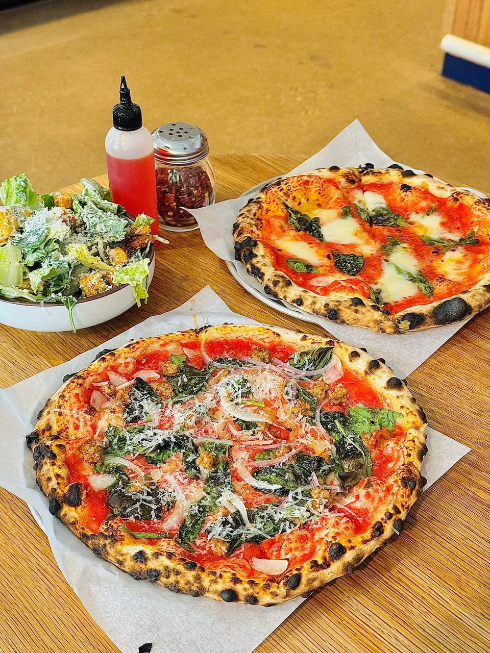 Two freshly baked Neapolitan-style pizzas with charred crusts, a Caesar salad, a bottle of chili oil, and a shaker of red pepper flakes on a wooden table.