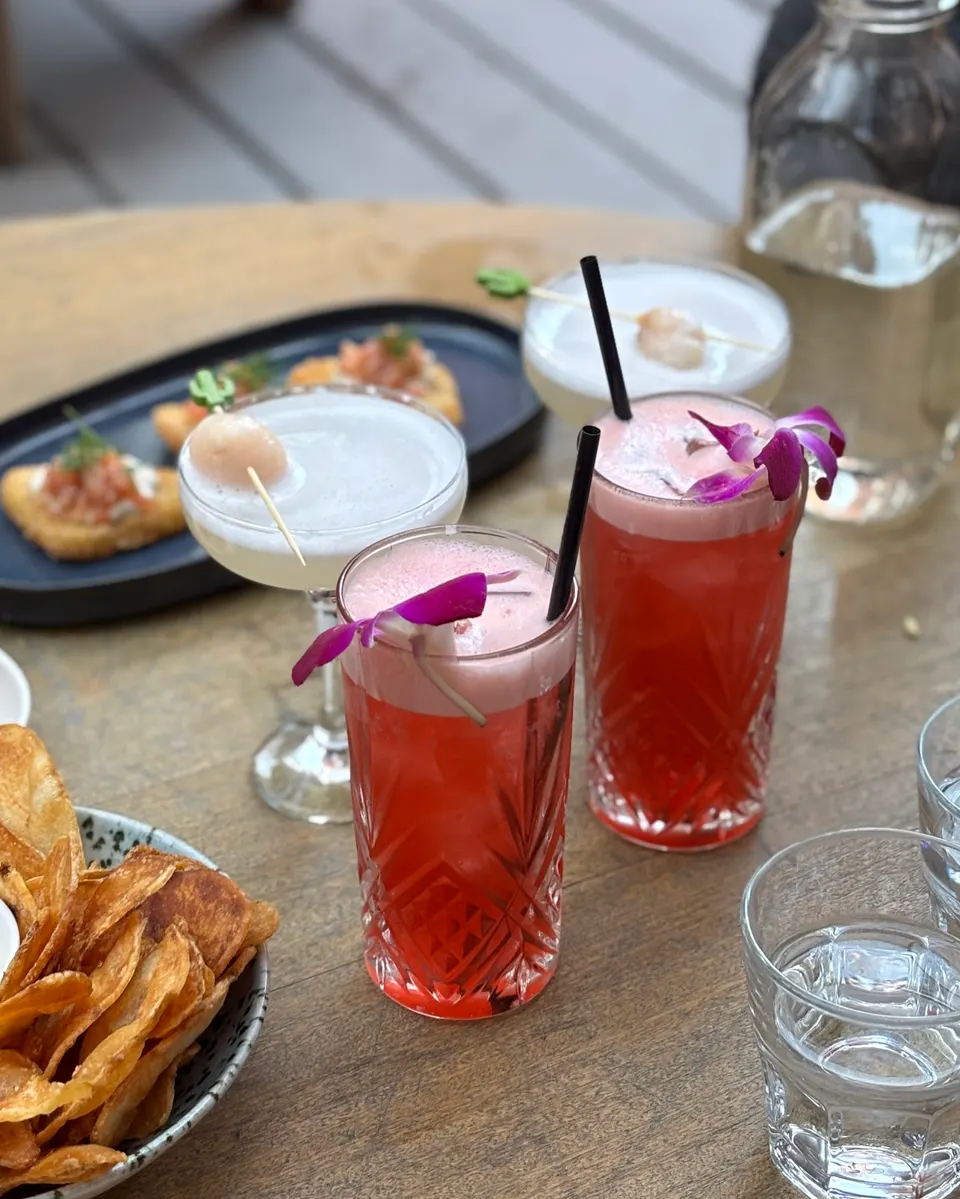 Four cocktails on a wooden table, including two tall red drinks with pink foam and purple flowers, and two white cocktails in coupe glasses garnished with lychees, alongside potato chips and bruschetta.