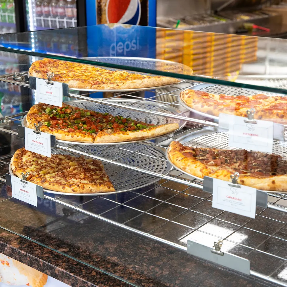 A multi-tiered metal display rack behind a glass counter showcasing several varieties of whole and sliced pizzas on round metal trays, including cheese, vegetarian, pepperoni, and Canadian styles.
