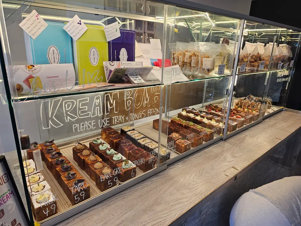 A glass display case filled with rows of square-shaped pastries, labeled with flavors like Taro, Earl Grey, Pistachio, and Creme Brulee, with gift boxes on the top shelf.