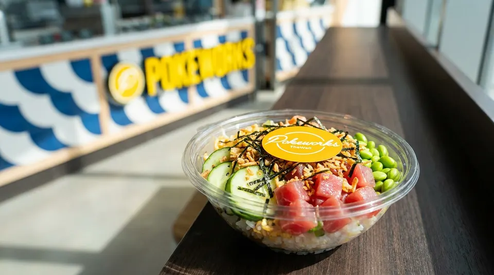 A fresh poke bowl with tuna, edamame, cucumber, and crispy onions in a clear plastic bowl on a wooden counter, with a blurry Pokeworks sign in the background.