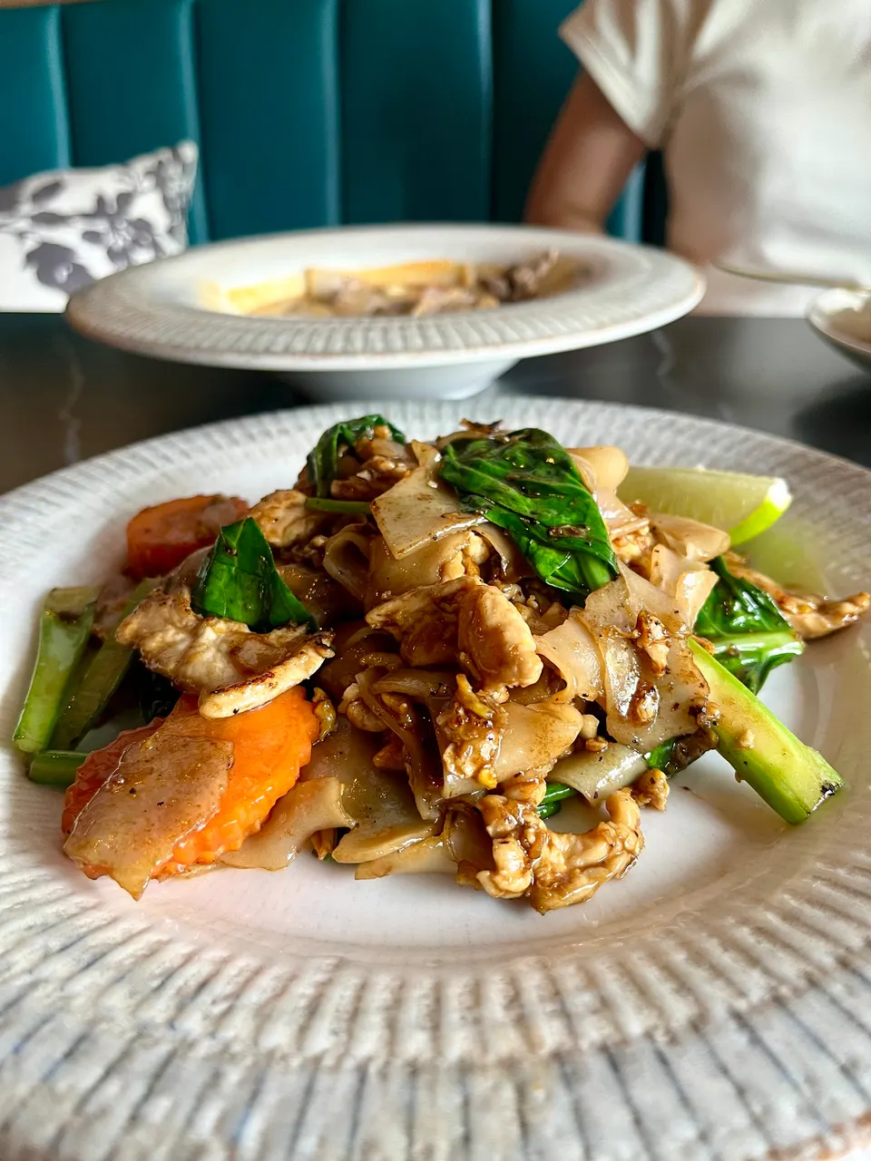 A close-up shot of a plate of Pad See Ew with wide rice noodles, chicken, Chinese broccoli, and carrots, served on a textured white plate with another dish blurred in the background.