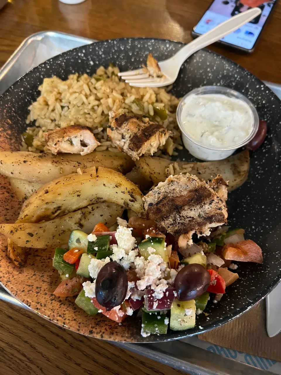 A Greek platter on a speckled plate featuring seasoned rice, roasted potato wedges, grilled chicken pieces, a small Greek salad with feta and olives, and a side of tzatziki sauce.