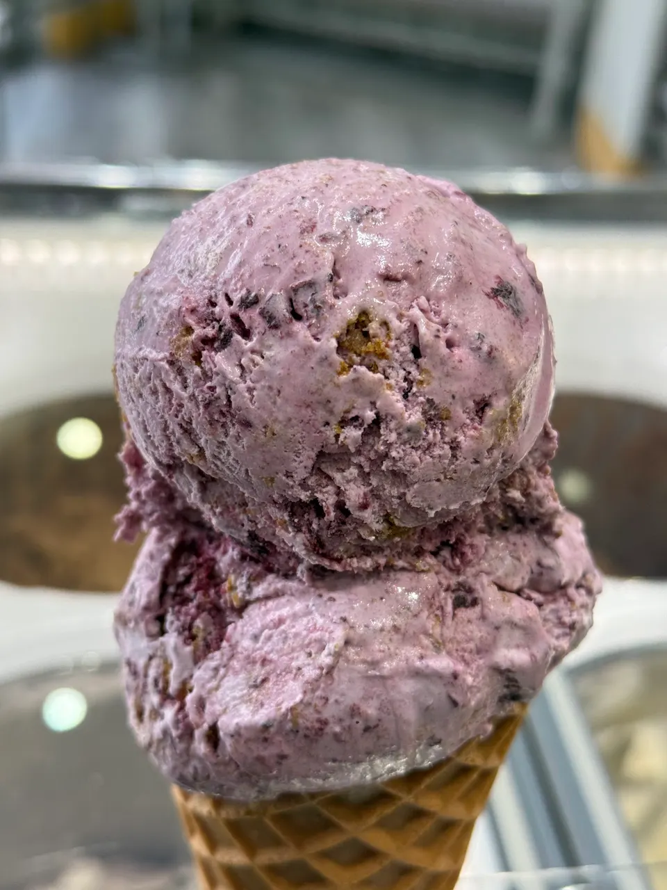 Two scoops of purple berry ice cream with cookie mix-ins stacked on a waffle cone, held up in front of a blurry ice cream display case.