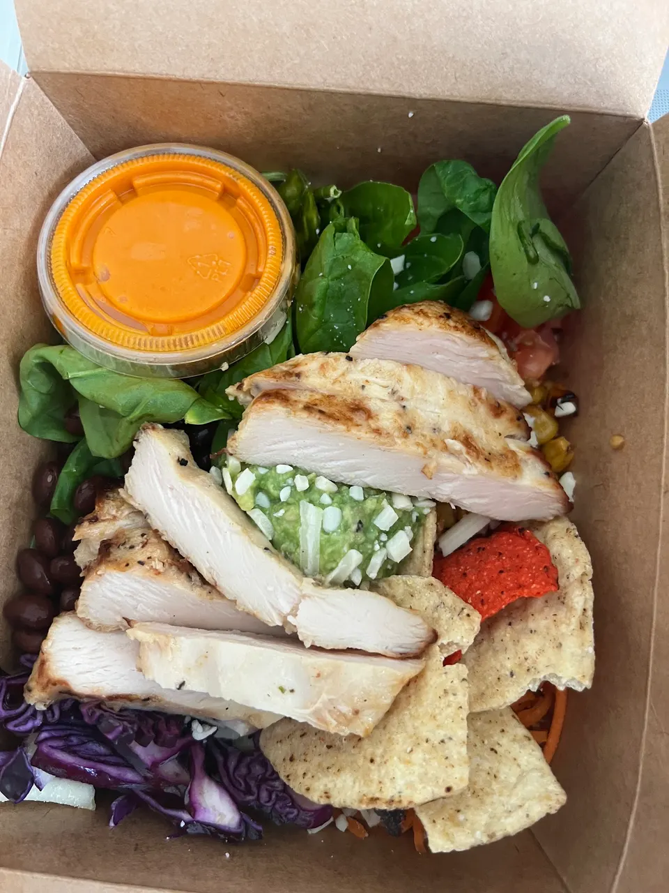 A takeout box containing a salad with grilled chicken breast slices, guacamole, tortilla chips, black beans, red cabbage, spinach, and a small container of orange dressing.