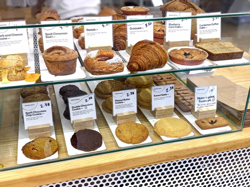 A glass display case at Robinson Bread bakery filled with fresh pastries, including croissants, danishes, cinnamon swirls, and various cookies with handwritten price tags.