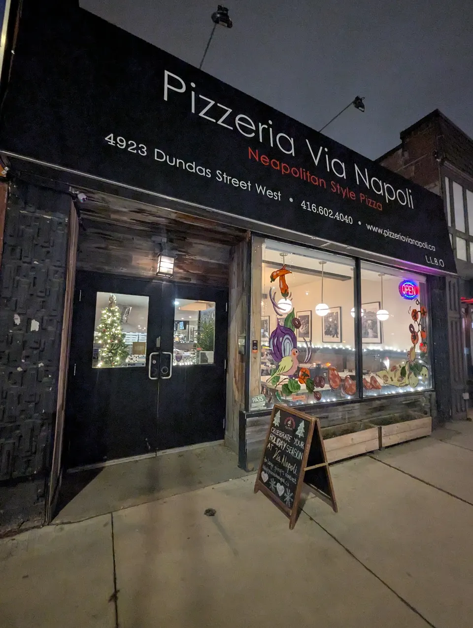 The storefront of Pizzeria Via Napoli at night, featuring a black sign with white and red text, a large window with colorful illustrations, and a wooden sandwich board on the sidewalk.