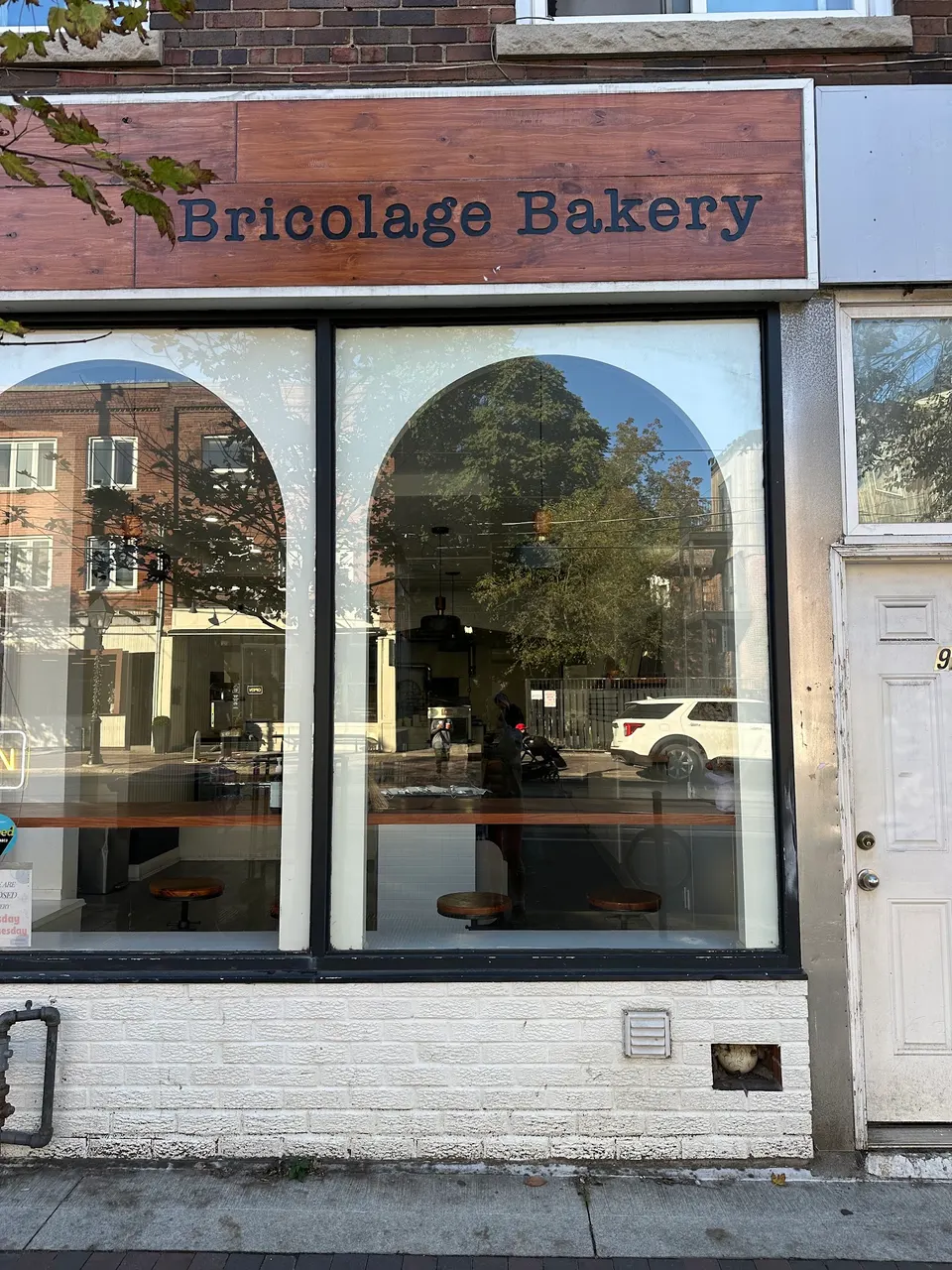 The storefront of Bricolage Bakery featuring a wooden sign with the bakery's name above large arched windows that reflect the street and trees outside.