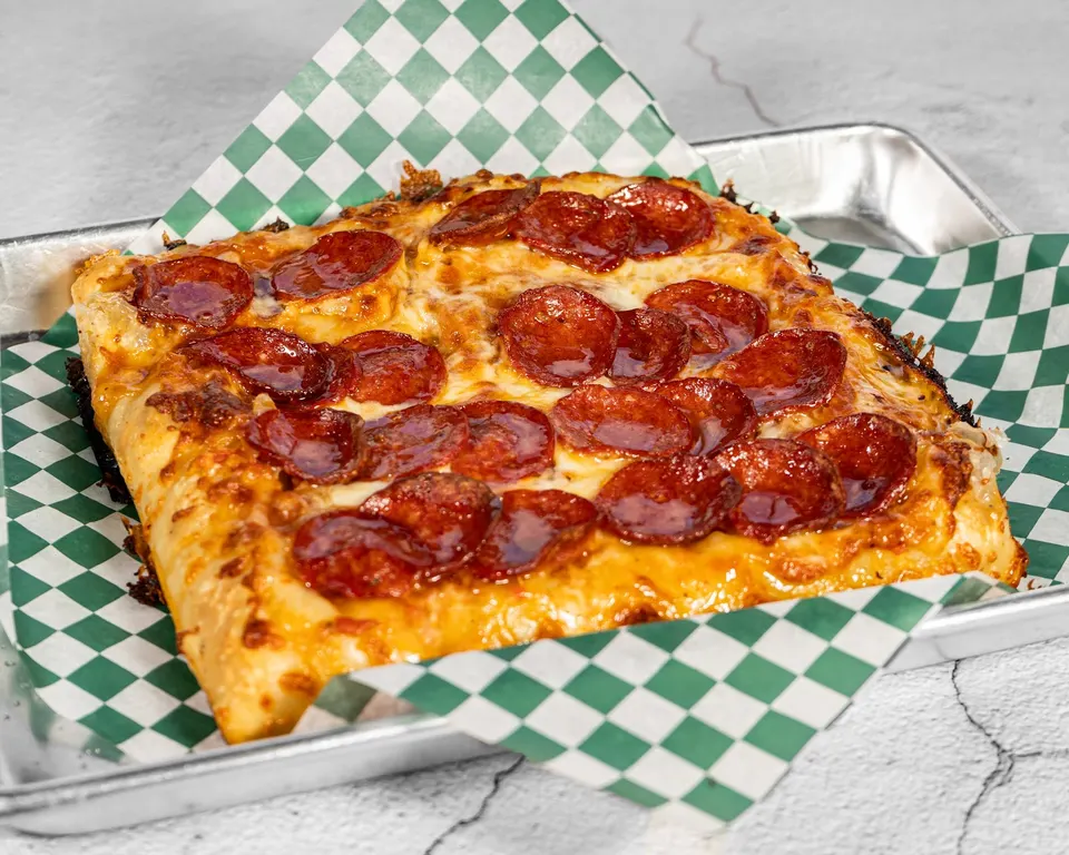 A square Detroit-style pepperoni pizza with a thick, crispy golden crust and melted cheese, served on green and white checkered paper inside a metal tray.