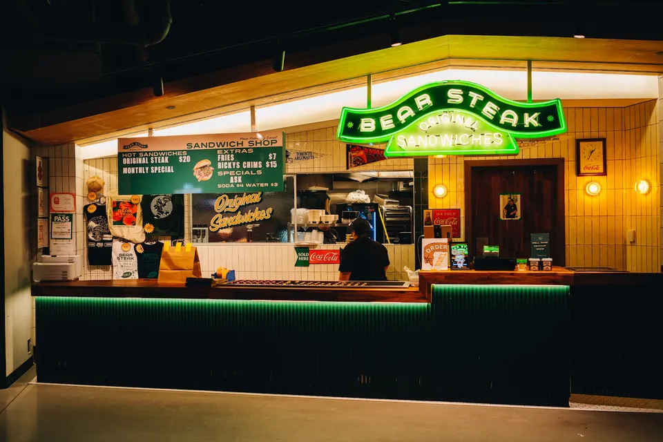 A sandwich shop counter with a glowing green neon sign reading 'BEAR STEAK ORIGINAL SANDWICHES' above a dark green fluted bar counter.