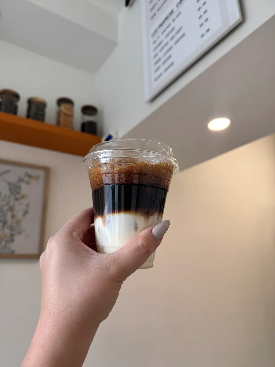 A hand holding a clear plastic cup containing a layered iced coffee drink with milk at the bottom and dark espresso on top, set against a cafe interior background.