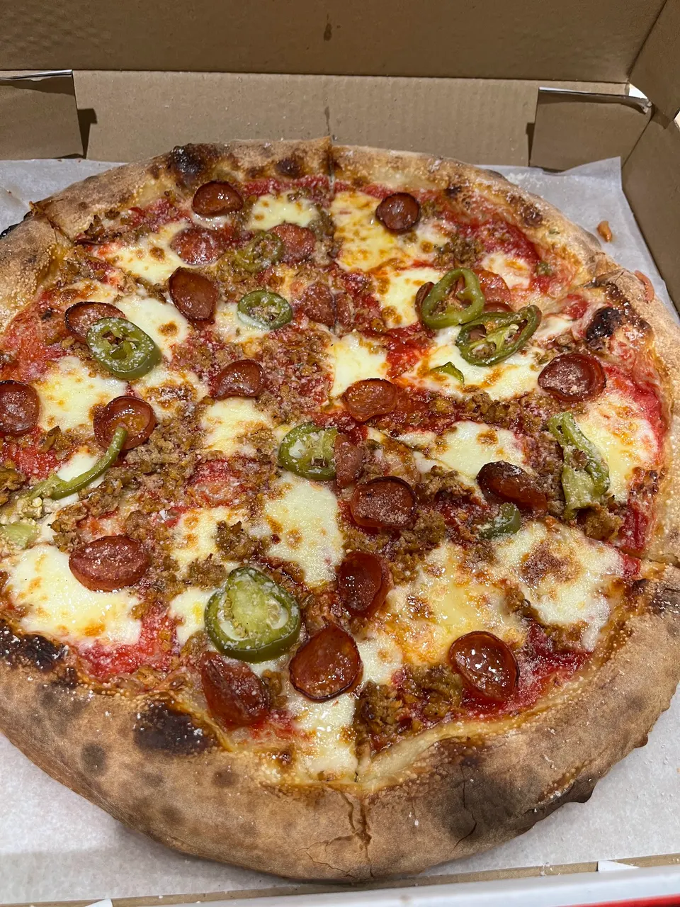 A freshly baked pizza in a cardboard box, topped with melted mozzarella cheese, pepperoni slices, crumbled sausage, and green jalapeño rings on a thin, charred crust.