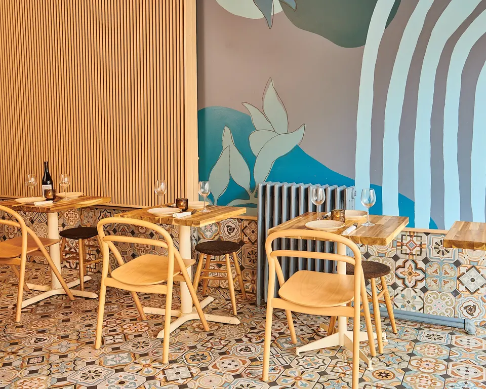 A cozy dining area at Levant Pizza featuring small wooden tables set with plates and wine glasses, light wood chairs, patterned floor tiles, and a colorful mural on the wall.