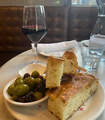 A plate of fresh focaccia bread and a small bowl of green and black olives served on a marble table next to a glass of red wine and a glass of water.