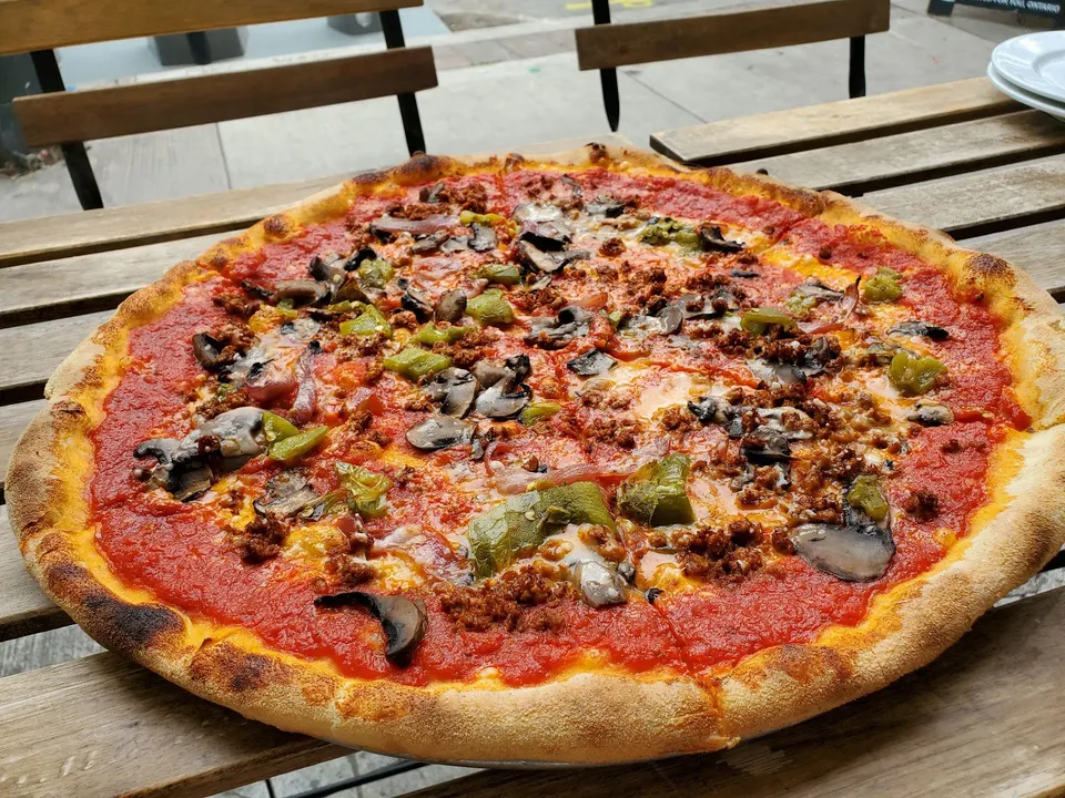 A large, thin-crust pizza topped with tomato sauce, melted cheese, mushrooms, green peppers, and ground meat, served on a wooden outdoor table.