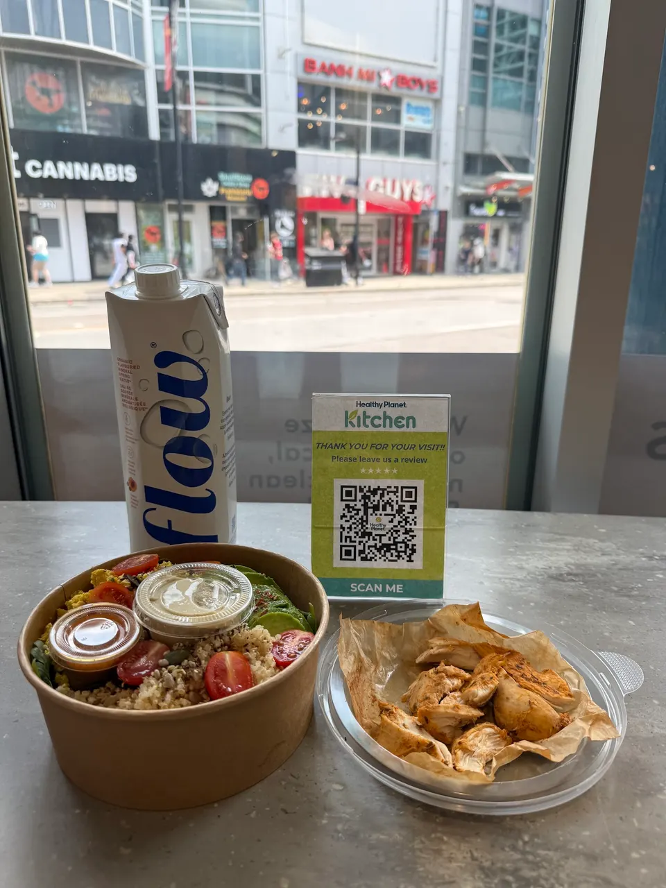 A healthy meal set on a table featuring a quinoa salad bowl with cherry tomatoes and avocado, a side of grilled chicken pieces, a carton of Flow water, and a Healthy Planet Kitchen QR code sign.