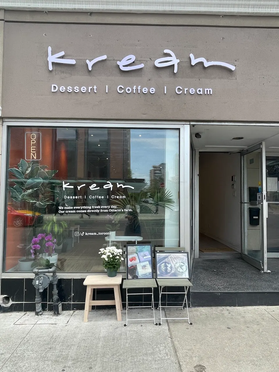 The storefront of Kream, a dessert, coffee, and cream shop, featuring a minimalist grey facade, large glass window with white lettering, and small outdoor seating with potted plants.