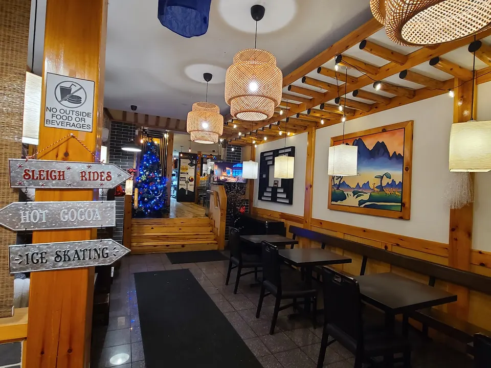 Cozy dining area of a Korean restaurant featuring wooden tables and chairs, woven pendant lights, a wooden trellis ceiling, and a decorative signpost pointing to winter activities.