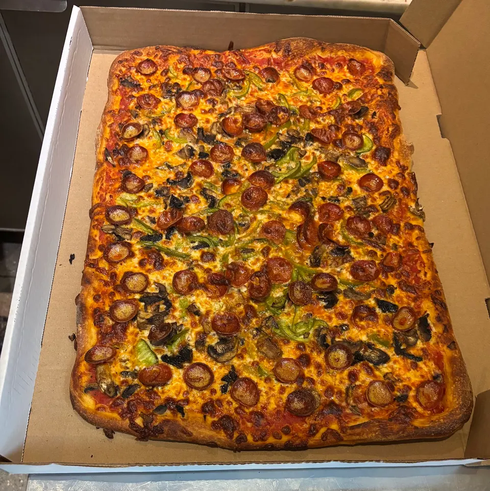 A large, rectangular party-sized pizza in a cardboard delivery box, topped with melted cheese, pepperoni, green bell peppers, and mushrooms.
