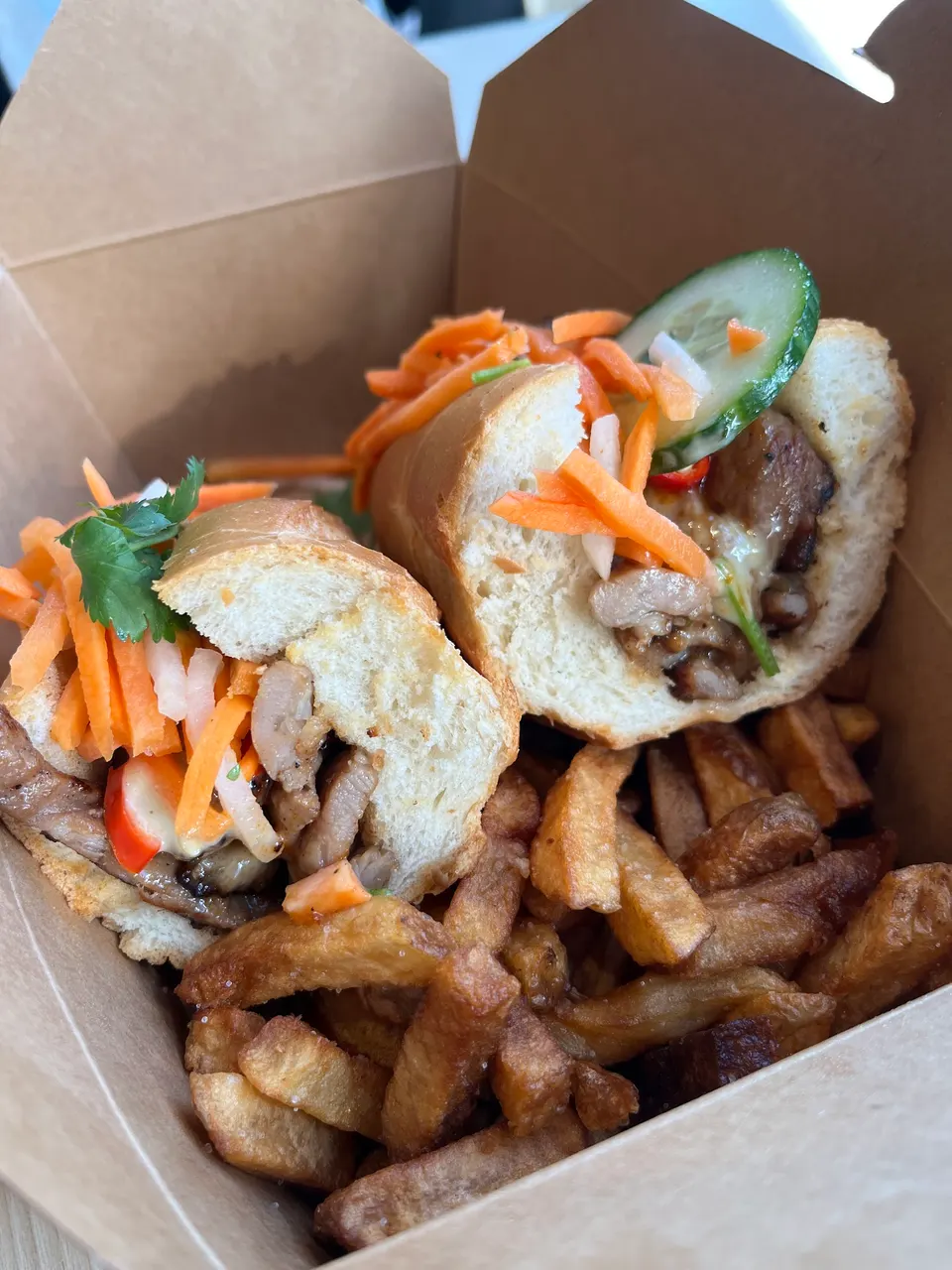 Two halves of a banh mi sandwich filled with grilled meat, pickled carrots, and cucumber, served with a side of thick-cut french fries in a cardboard takeout box.