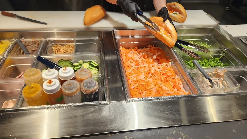 A food prep station at And Banh Mi showing a worker in black gloves using tongs to fill a crusty baguette with pickled carrots and daikon from a large metal tray.