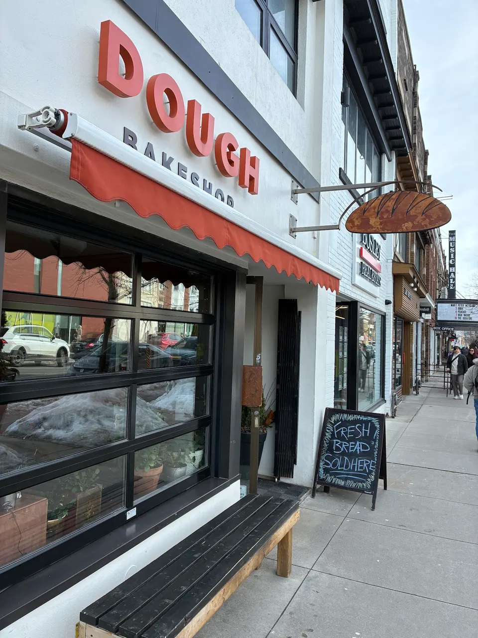 Exterior view of Dough Bakeshop featuring a large orange sign, a matching orange awning, a wooden bench, and a chalkboard sign reading 'Fresh Bread Sold Here' on the sidewalk.