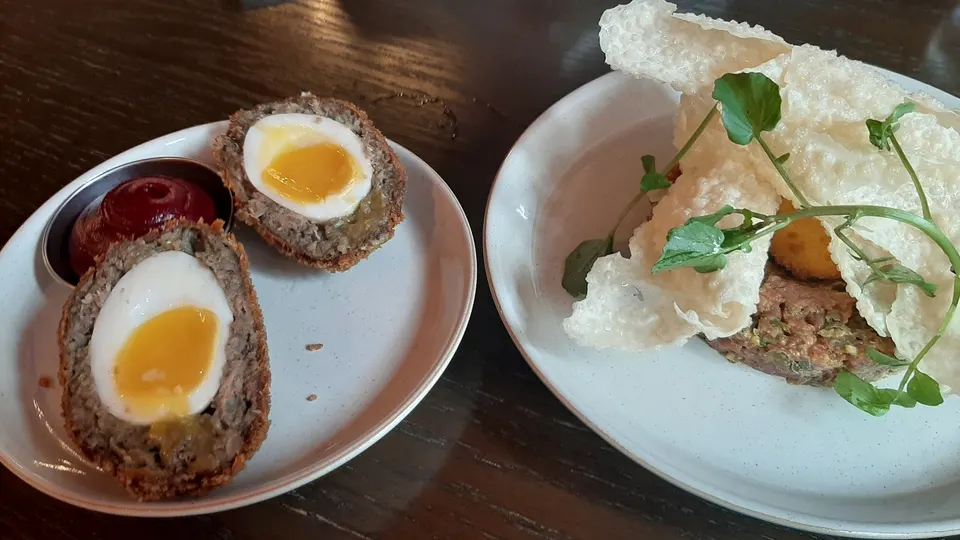 Two plates of food on a dark wooden table. The left plate has two halved Scotch eggs with runny yolks and a side of red sauce. The right plate features a tartare dish topped with large white crispy crackers and green herbs.