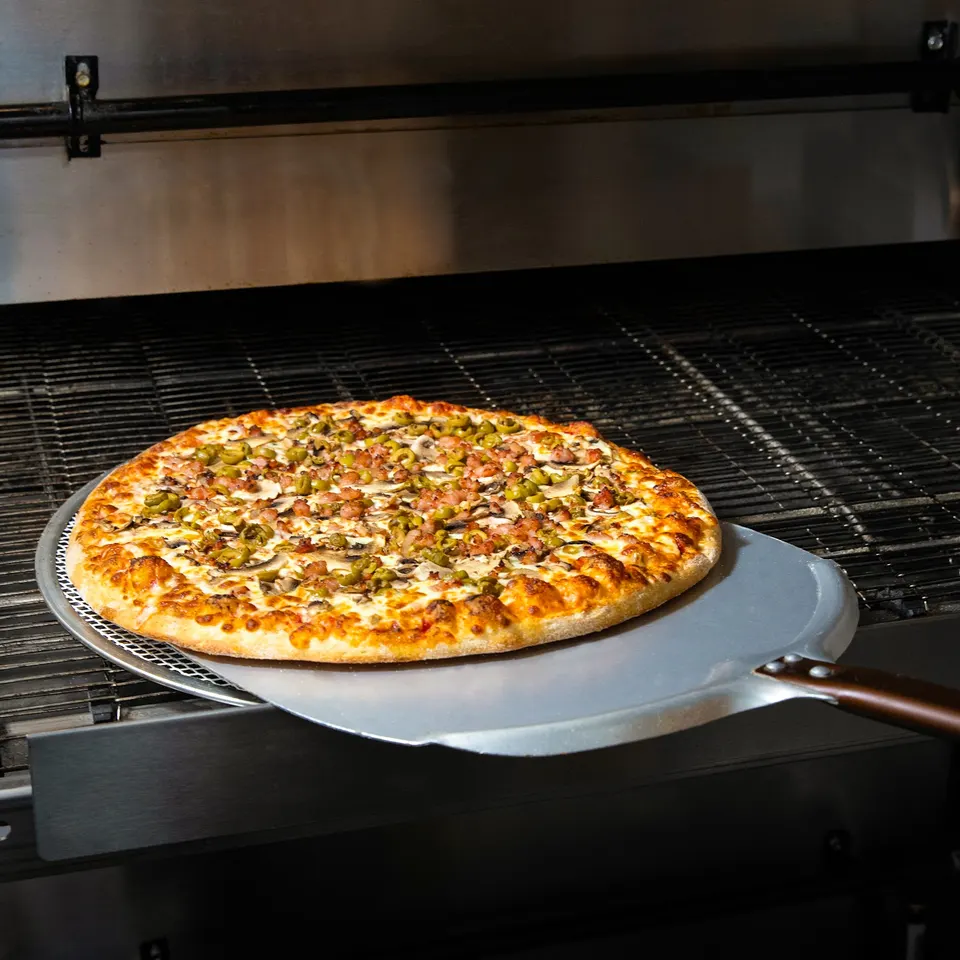 A freshly baked pizza topped with green olives, mushrooms, and bacon is pulled from a commercial conveyor oven using a metal pizza peel.