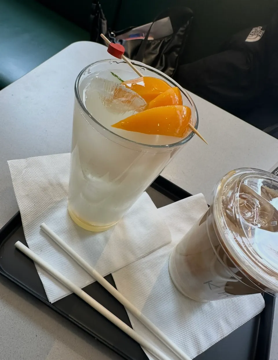 A tall glass of iced lemonade garnished with skewered peach slices sits on a black tray next to an iced coffee in a plastic cup with two white straws.