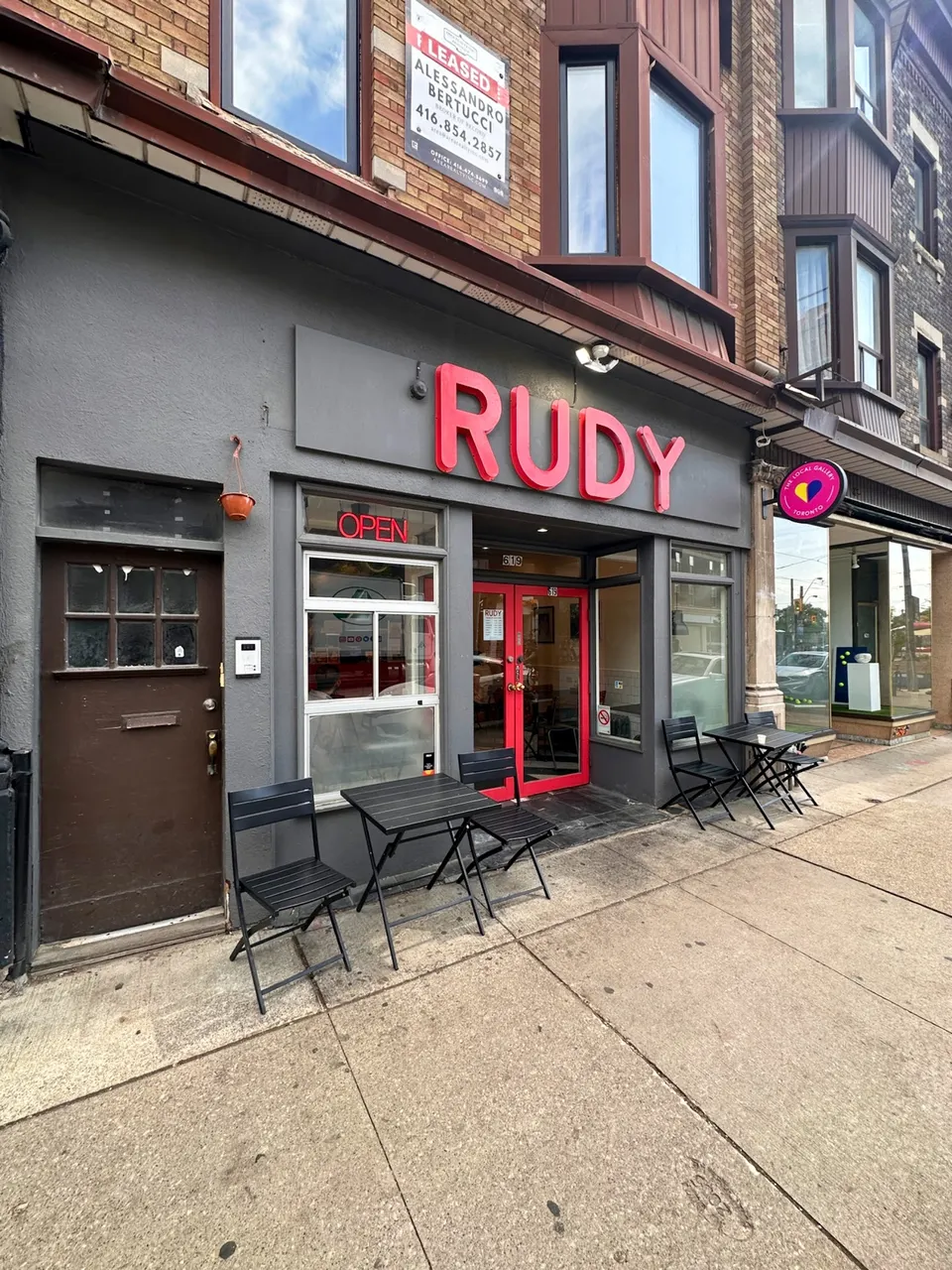 Exterior storefront of RUDY restaurant with a grey facade, large red sign, red-framed glass door, and two small black outdoor tables with chairs on the sidewalk.