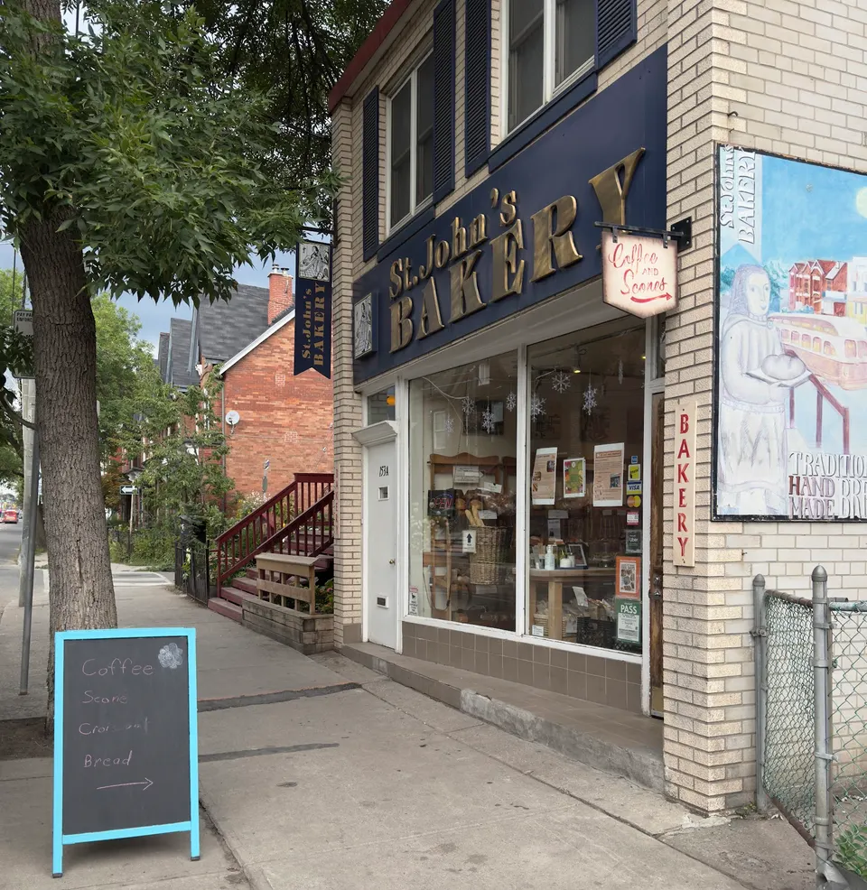Exterior view of St. John's Bakery, a brick building with a blue storefront sign, large display windows, a chalkboard sign on the sidewalk, and a painted mural on the side wall.