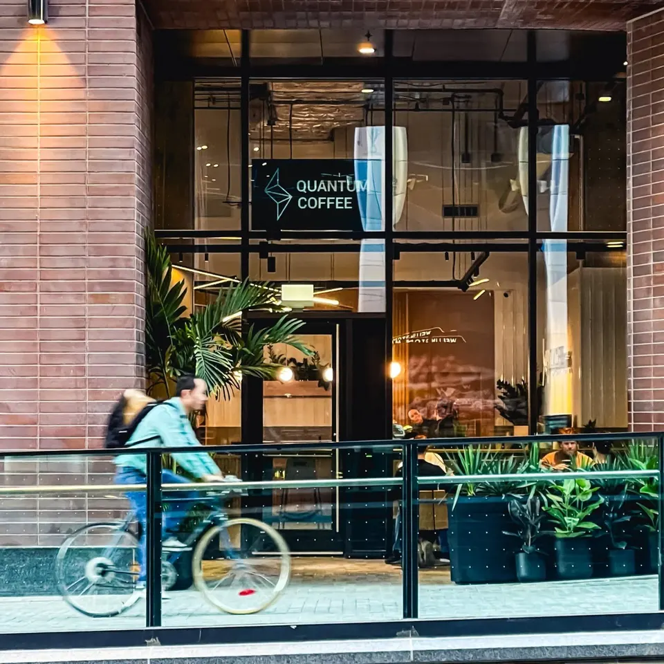 Exterior view of Quantum Coffee shop with a black sign displaying its name and logo. The shop has large glass windows reflecting the surroundings. A person on a bicycle is riding past in the foreground, and there are potted plants along the railing.