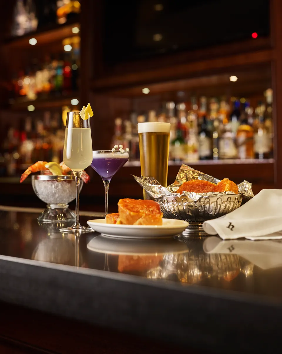 A selection of cocktails, a tall glass of beer, shrimp cocktail, and warm bread rolls served on a polished bar counter at Hy's Steakhouse & Cocktail Bar.