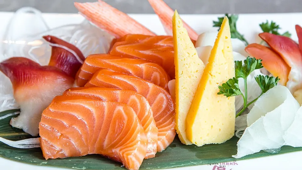 A close-up of a Japanese sashimi platter featuring thick slices of fresh salmon, tamago egg omelet, surf clam, and crab sticks arranged on a green leaf.