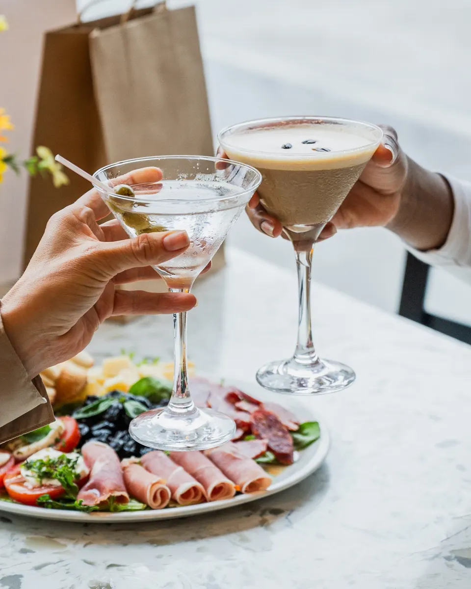 Two people toast with cocktails over a plate of Italian antipasto on a marble table. One holds a classic martini with an olive, and the other holds an espresso martini.