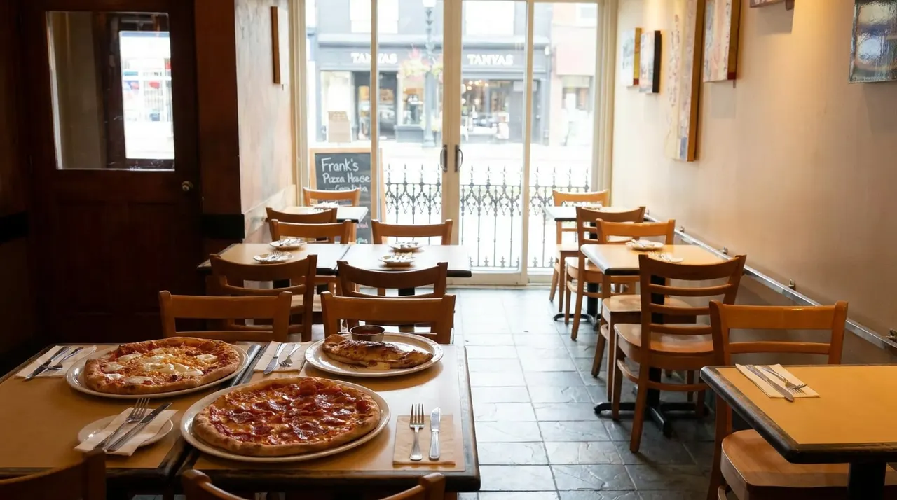 An empty dining room at Frank's Pizza House with wooden tables and chairs, featuring freshly baked pizzas and a calzone on the foreground tables near a glass door leading outside.
