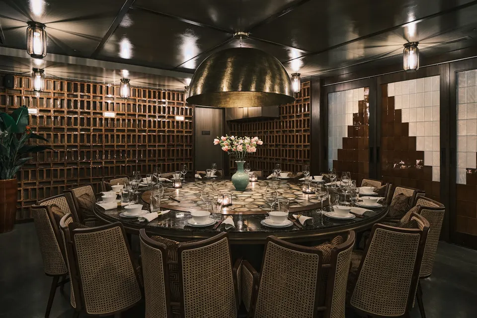 A large round dining table with a lazy Susan, set for a group dinner with elegant tableware, surrounded by woven-back chairs under a large brass dome pendant light in a moody, upscale dining room.