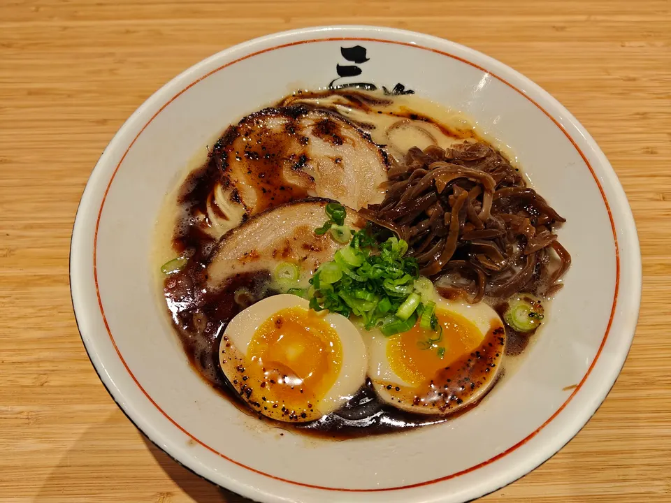 A bowl of tonkotsu ramen with two slices of chashu pork, a soft-boiled egg cut in half, black garlic oil, wood ear mushrooms, and chopped green onions, served in a white bowl on a wooden table.