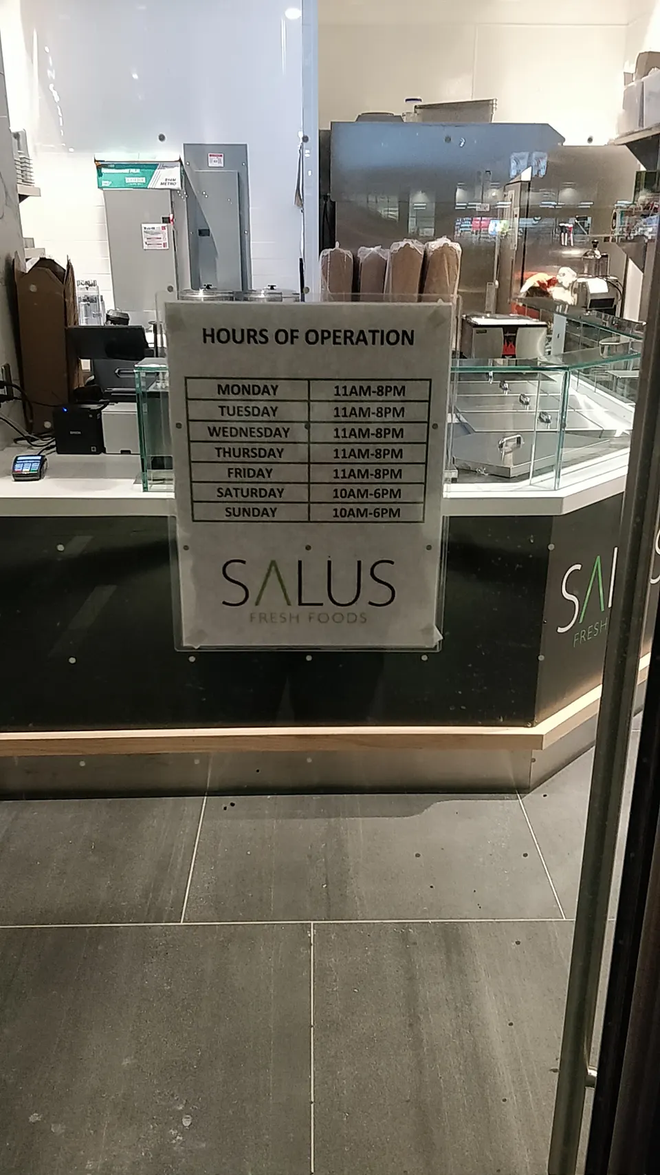 A sign posted on a glass partition at Salus Fresh Foods displays the hours of operation, showing Monday through Friday from 11 AM to 8 PM, and Saturday and Sunday from 10 AM to 6 PM.