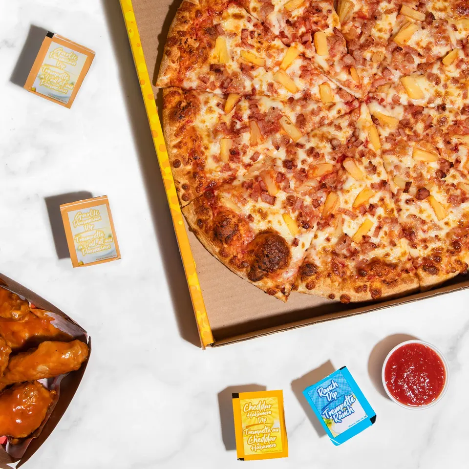 An overhead view of a large Hawaiian pizza with pineapple and ham in a cardboard box, served with chicken wings, marinara sauce, and various dipping sauce packets on a white marble surface.