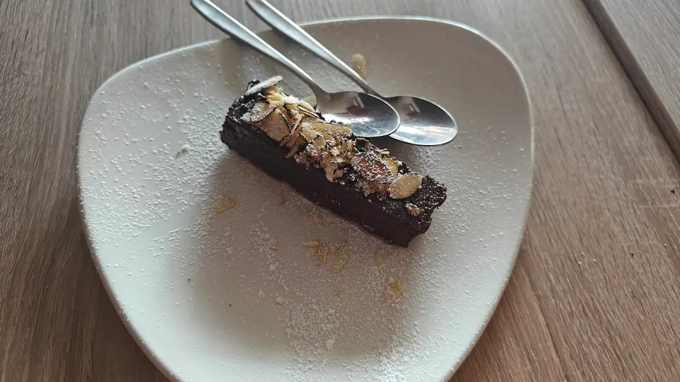 A rectangular slice of dark chocolate cake topped with sliced almonds and powdered sugar, served on a white triangular plate with two small spoons.