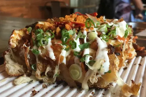 A close-up shot of a thick-crust Detroit-style pizza slice loaded with toppings, drizzled with white sauce, and garnished with chopped green onions on a white ribbed serving tray.