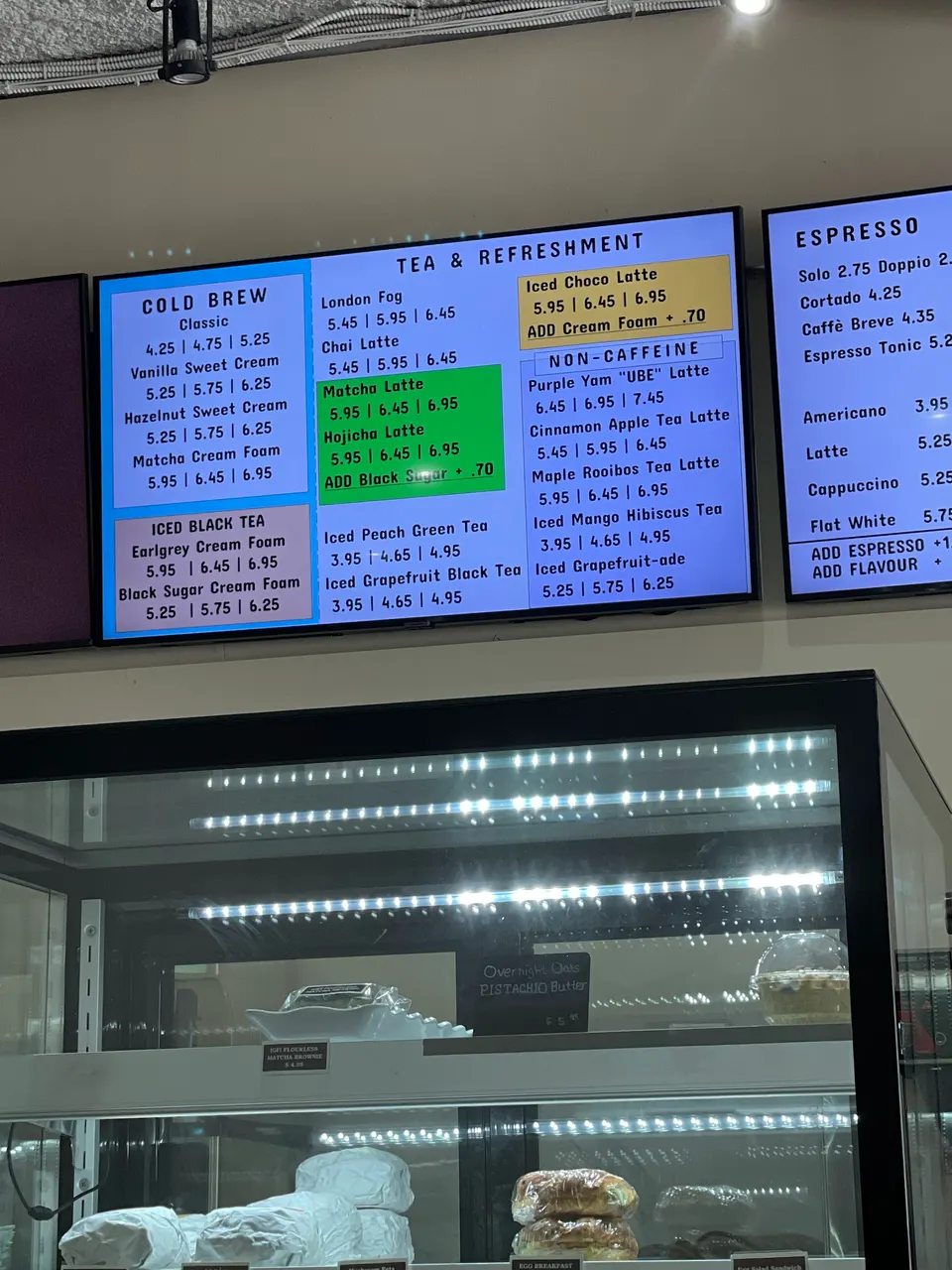 A digital menu board displaying cold brew, tea, and espresso options above a glass display case filled with wrapped baked goods and pastries.