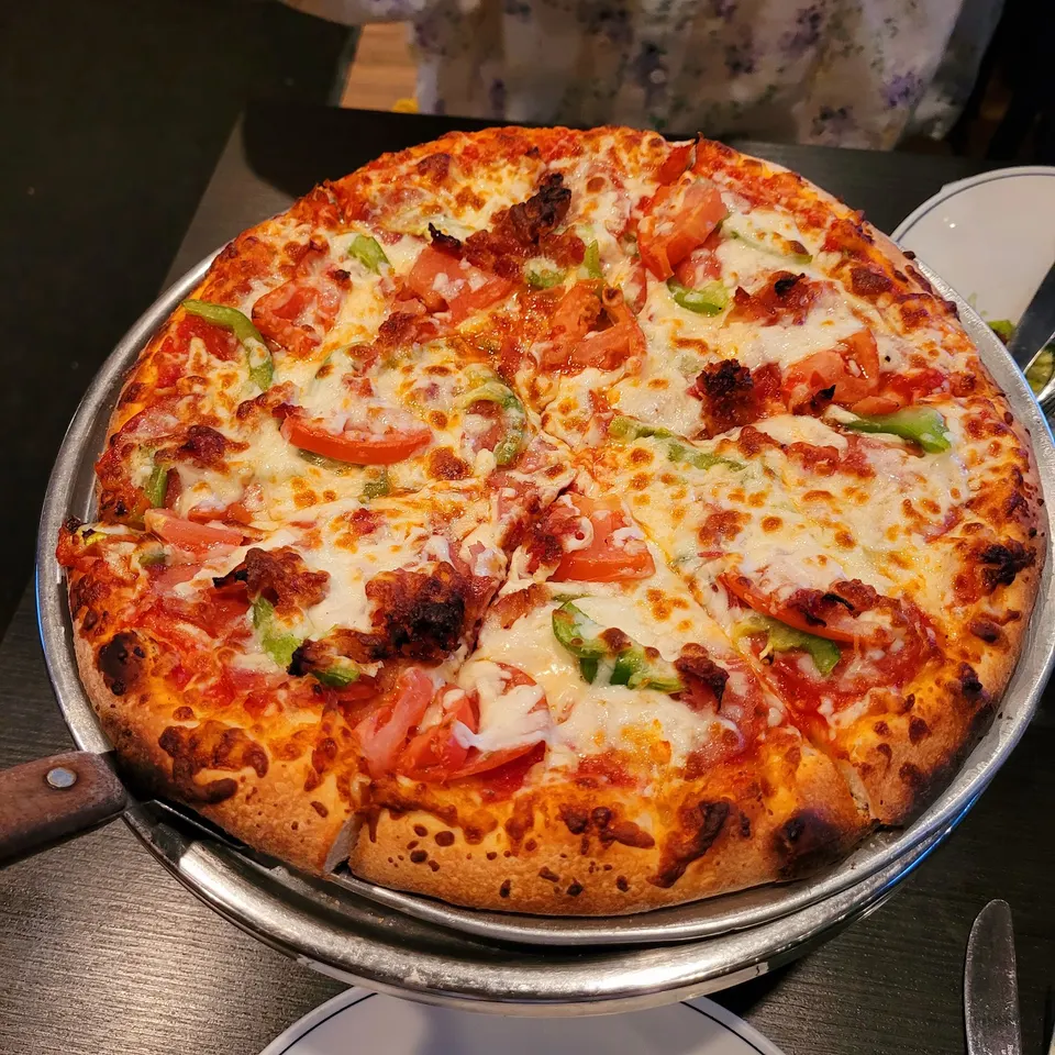 A freshly baked whole pizza with thick crust, melted mozzarella cheese, sliced tomatoes, and green bell peppers, served on a metal tray at a restaurant table.