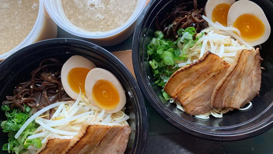 Two black takeout bowls filled with ramen ingredients including sliced pork belly, soft-boiled eggs, bean sprouts, green onions, and wood ear mushrooms, next to two containers of broth.