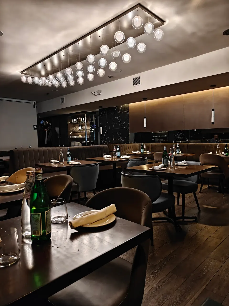 An elegant dining room at KHAZANA Toronto featuring dark wood tables set with water bottles and glasses, comfortable upholstered chairs, and a striking modern chandelier with multiple glowing glass globes.