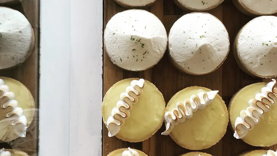 A top-down view of neatly arranged tarts on a wooden tray, featuring some with high mounds of white whipped topping and others with smooth yellow filling decorated with a wave of toasted meringue.