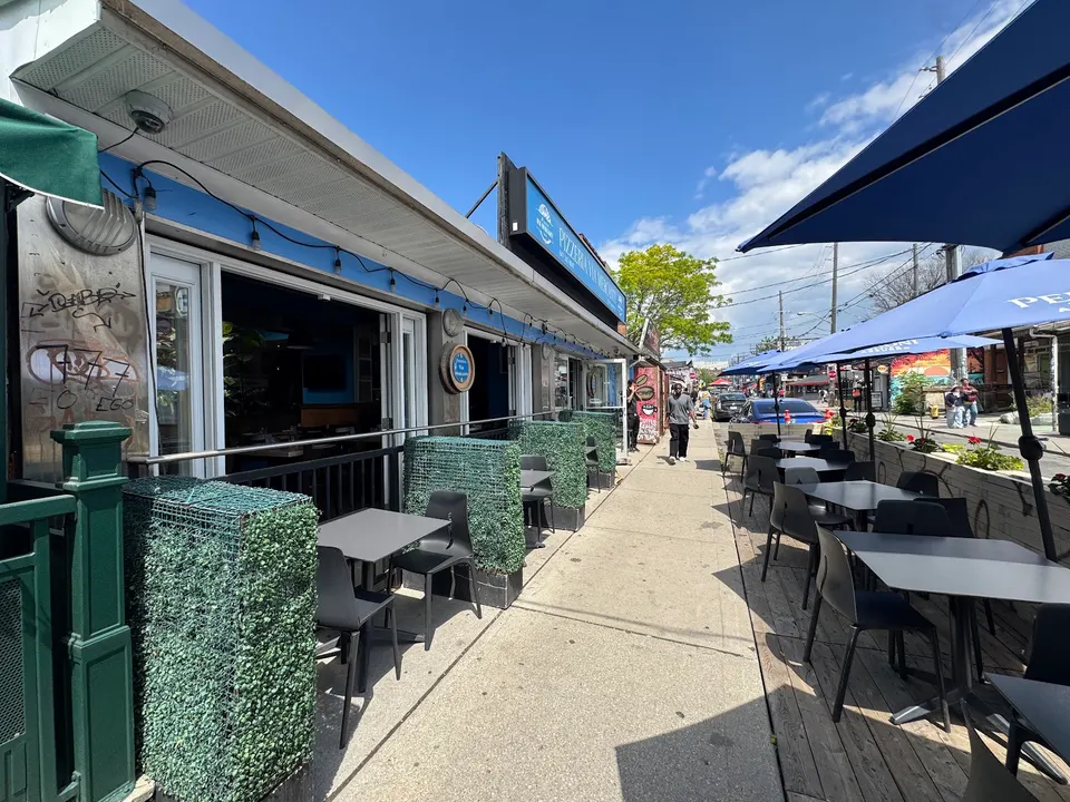 Outdoor patio seating of Pizzeria Via Mercanti along a sunny sidewalk, featuring dark tables and chairs separated by green privacy hedges under blue umbrellas.