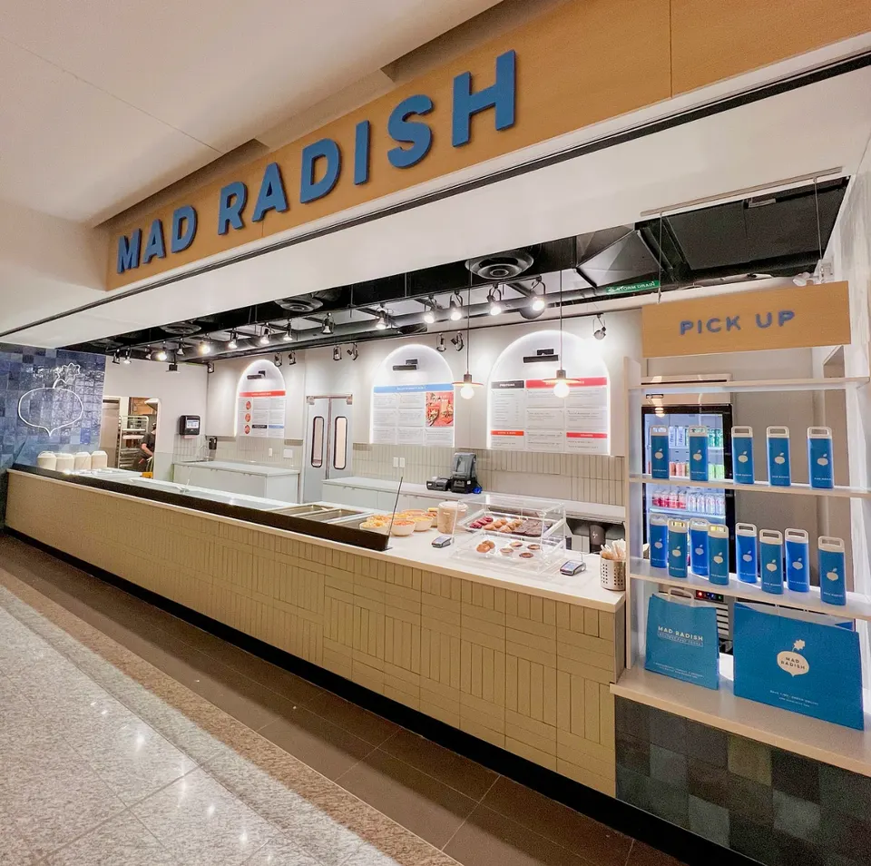 The modern counter and storefront of Mad Radish, featuring a light wood service counter, menu boards on the back wall, a pick-up station with branded water bottles, and a large blue sign overhead.