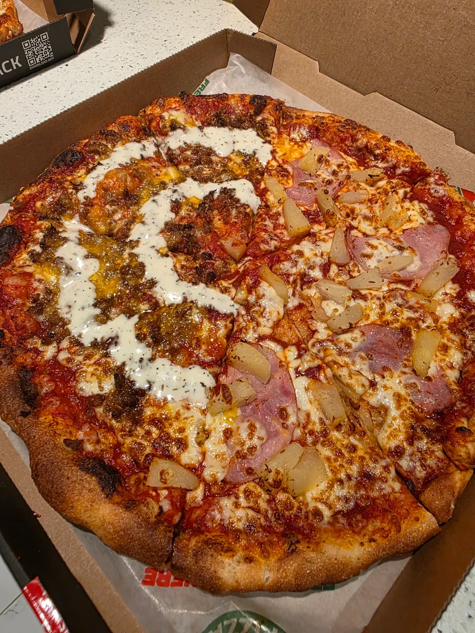 A whole pizza in a cardboard delivery box, split half-and-half with one side featuring ham and pineapple chunks, and the other side topped with ground meat and a white sauce drizzle.